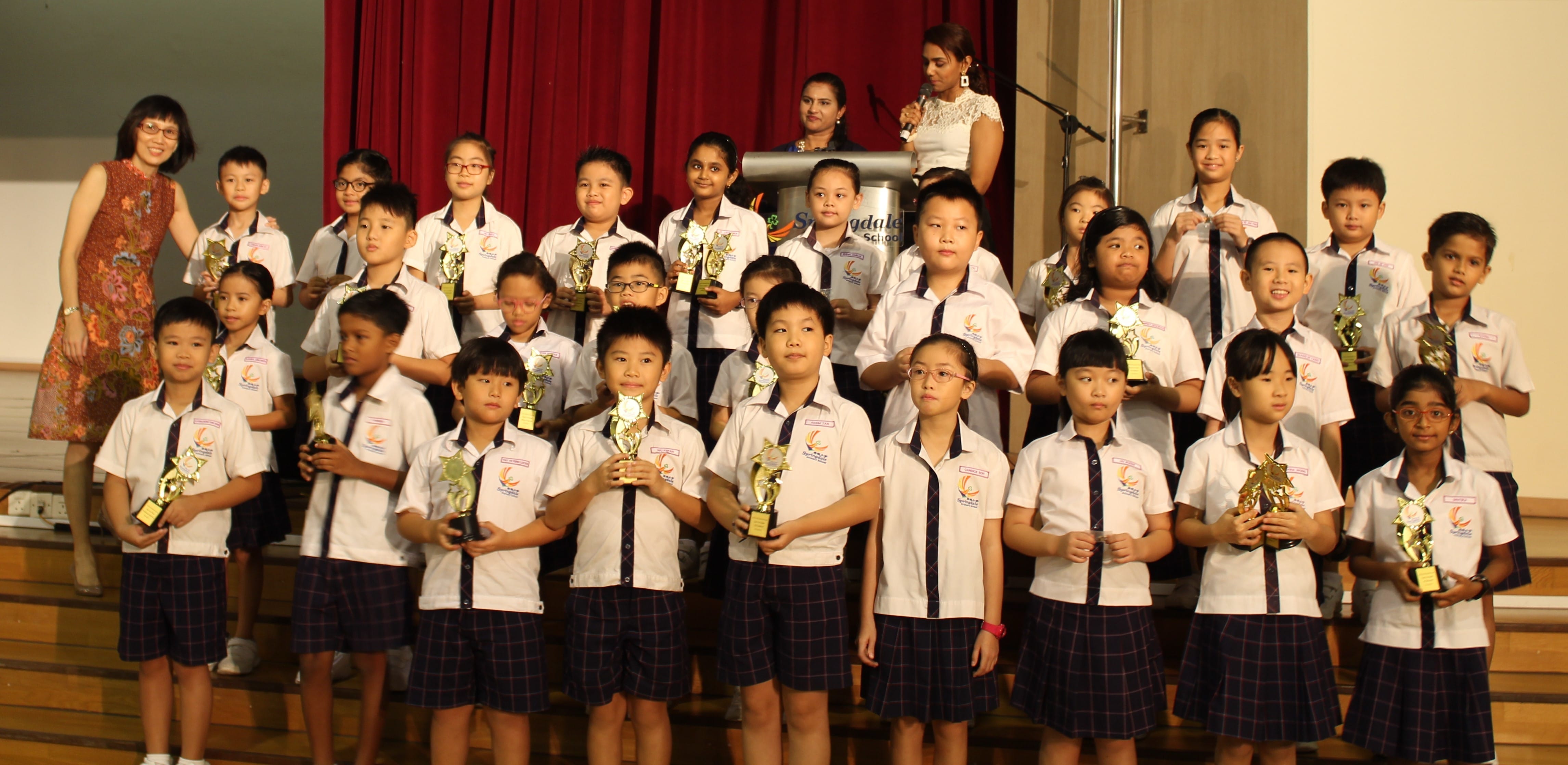A group of young students in school uniforms hold gold trophies on a stage with a red curtain backdrop.
